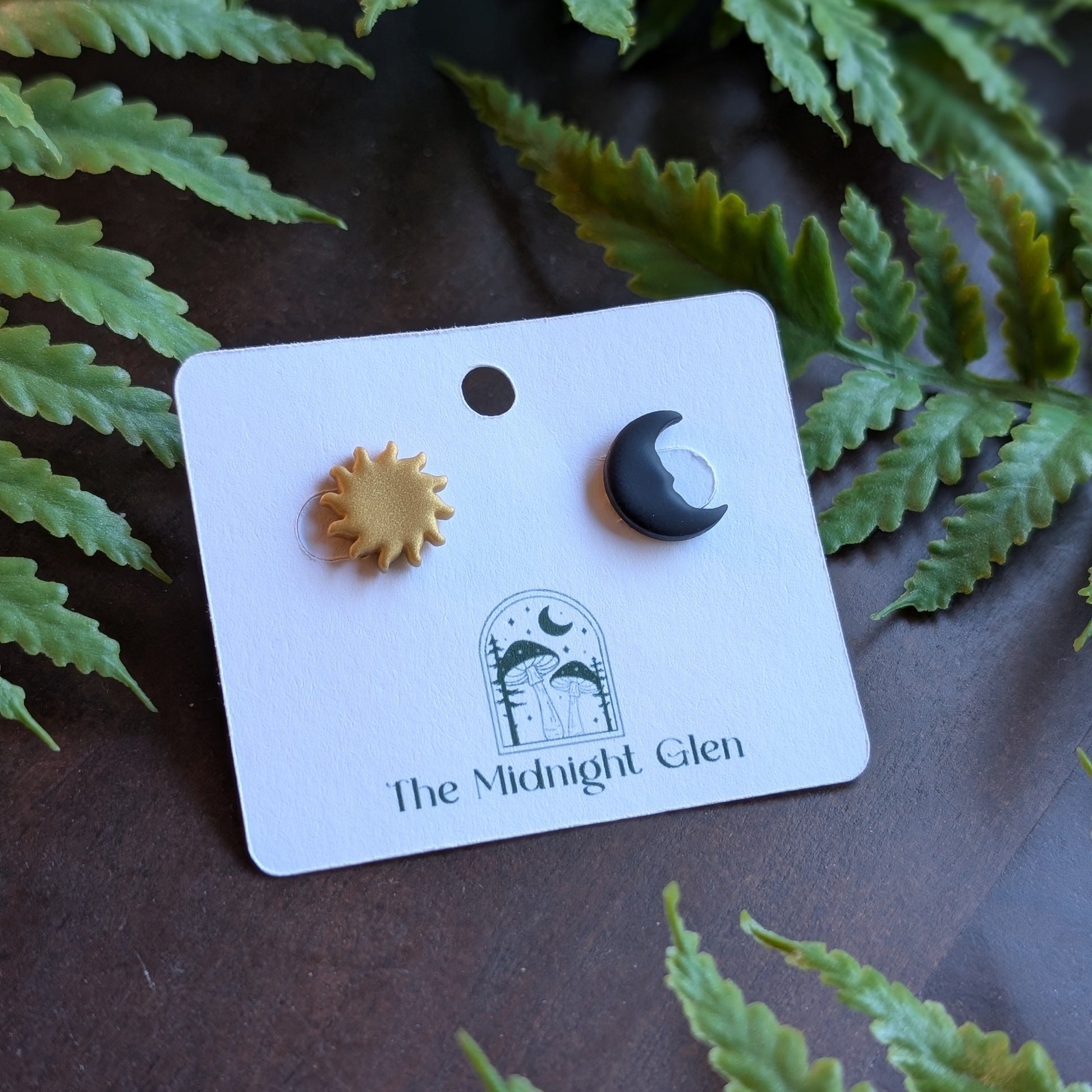Sun and moon earrings on a 'The Midnight Glen' branded card with fern leaves in the background