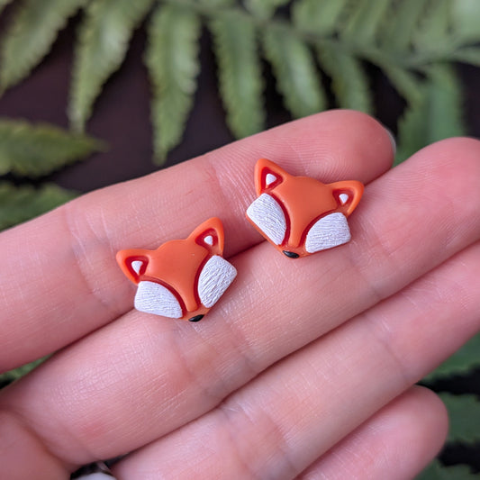 A pair of small fox-head stud earrings held on a person's fingers, with blurred green fern leaves in the background.