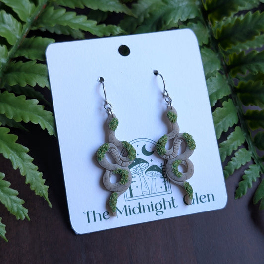 Pair of snake-shaped earrings on a 'The Midnight Glen' branded card with ferns in the background.