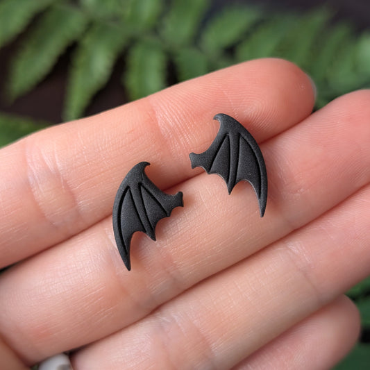 Black bat-wing shaped stud earrings held on a person's fingers with a blurred green plant background.
