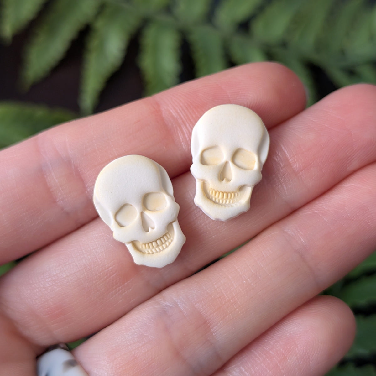 Two small skull-shaped stud earrings held on a person's fingers, with blurred green leaves in the background.
