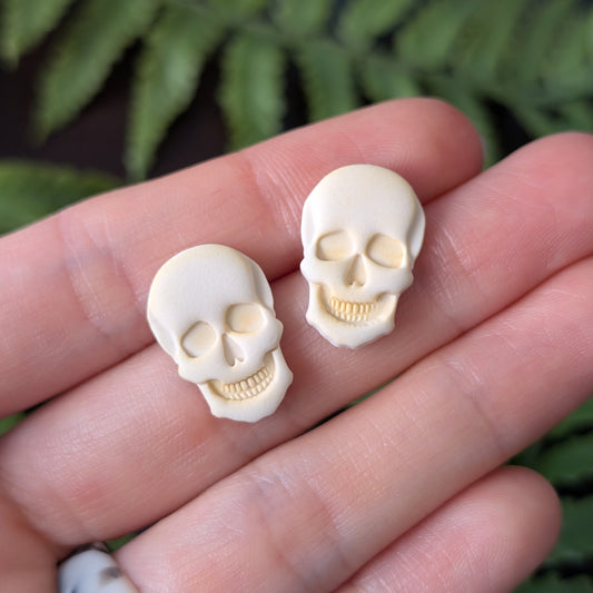 Two small skull-shaped stud earrings held on a person's fingers, with blurred green leaves in the background.